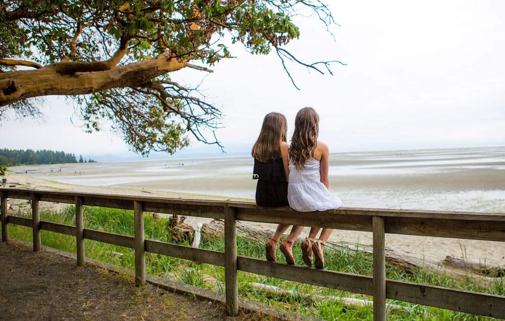 Two girls with long brown hair, left side girl wearing black dress and right side girl wearing white dress, sitting side by side on top of a wooden fence looking out to a beach with a large arbutus tree to the left