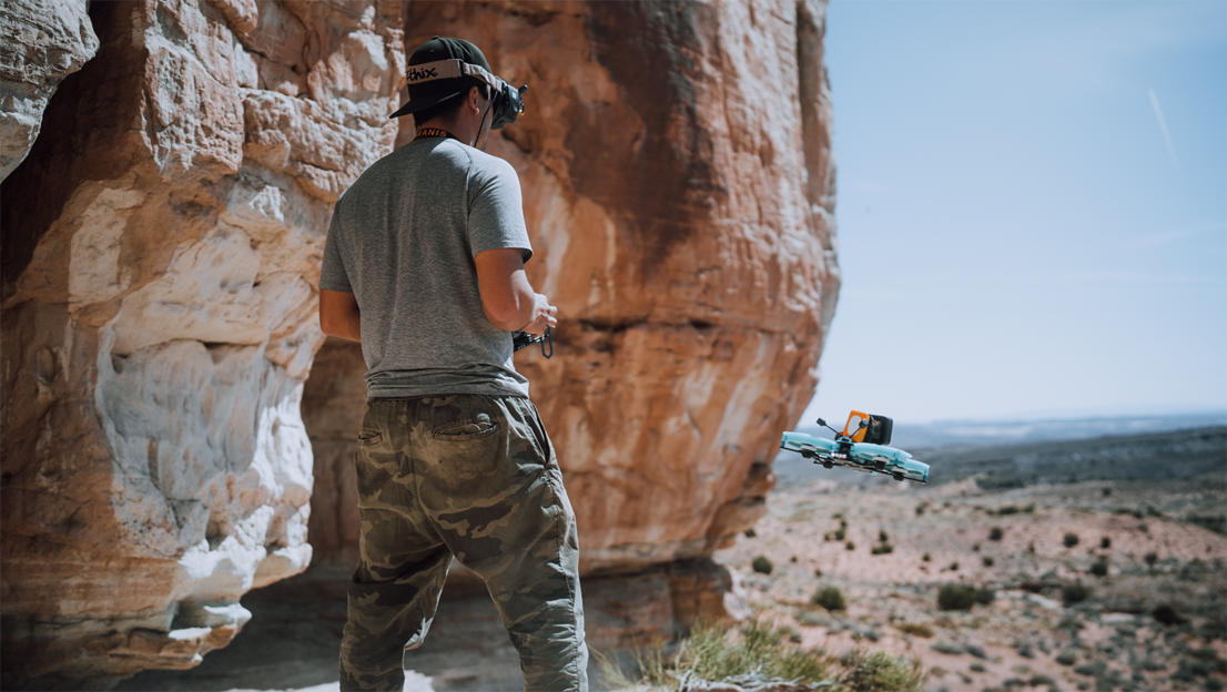 Man wearing a grey shirt and khaki pants with black hat backwards flying drone in valley of desert